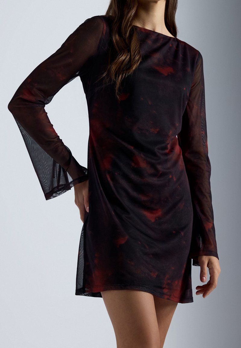 Femme portant une robe mini noire et rouge foncé à motif, transparente, manches longues et silhouette ajustée, sur un fond clair uni.