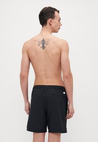 Calções de banho pretos com um cós elástico, um bolso traseiro e um detalhe de logotipo circular. Usados por uma pessoa com uma tatuagem nas costas.