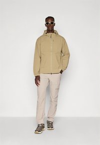 Vaaleanbeige tuulitakki, jossa on etuvetoketju, suuri huppu ja joustavat hihansuut, yhdistettynä vaaleanruskeisiin housuihin ja harmaisiin vaelluskenkiin, joissa on keltaiset yksityiskohdat.