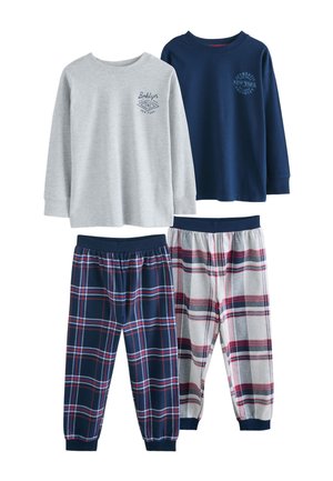 Deux garçons en pantalon de pyjama à carreaux et hauts à manches longues se tiennent dans un salon, l’un pieds nus, l’autre portant des chaussons bleu marine.