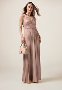 Maxikleid in zartem Pink mit plissierter Rockpartie, verziertem Oberteil und tiefem V-Ausschnitt. Akzentuiert mit einer cremefarbenen Handtasche. Weiche, fließende Stoffe.