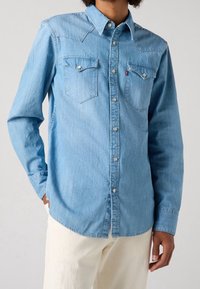 Vaaleansininen farkkupaita, jossa on levitetty kaulus, kaksi rintataskua läpillä, hopeiset napit ja kaartuva helma. Yhdistetty kermaan sävyisiin housuihin.