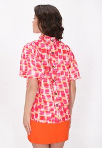 Kortærmet bluse i pink og orange penselstræk mønster, med en flæset halsudskæring og en knaplukning bagpå med brune knapper.