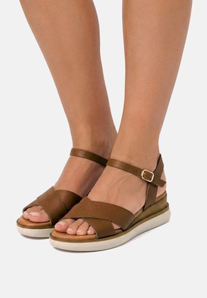 Füße mit braunen Leder-Open-Toe-Sandalen mit Knöchelriemen und weißen gepolsterten Sohlen vor einem schlichten hellen Hintergrund.