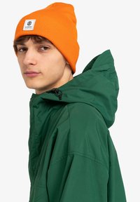 Giacca verde con cappuccio, colletto alto e cordoncino, abbinata a un berretto arancione a coste con una toppa del logo. Texture del tessuto liscia.
