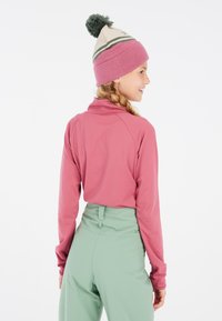 Haut de sport à manches longues, rose, avec un col montant, associé à un pantalon taille haute vert menthe. La mannequin porte un bonnet rose et crème avec un pompon.