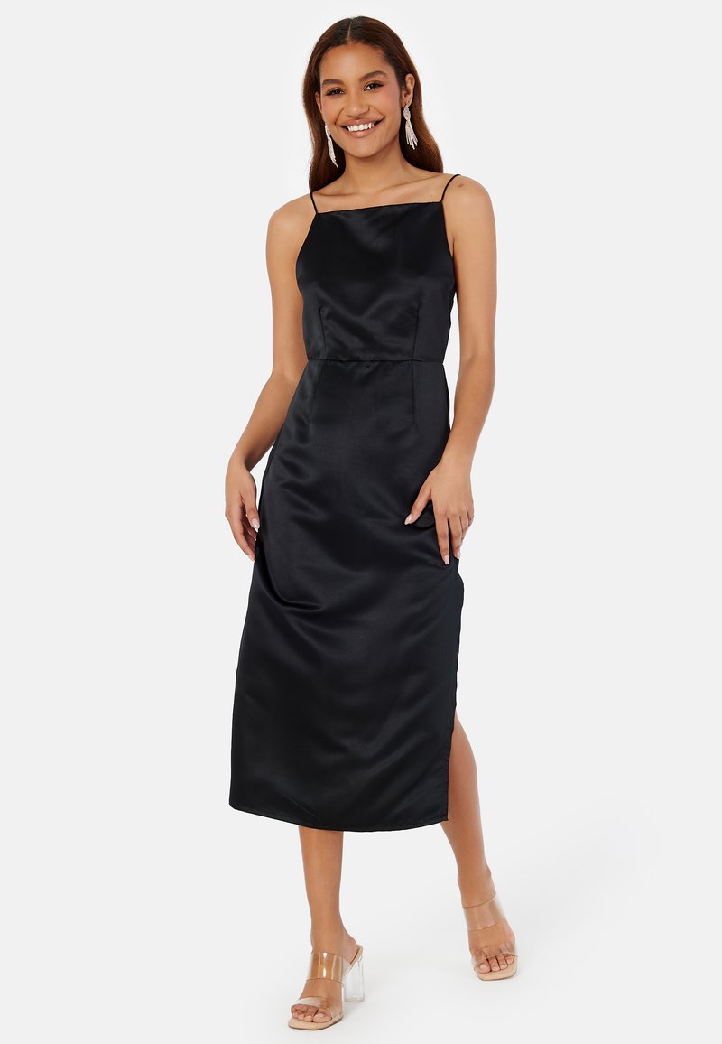 Abito midi in satin nero con scollatura quadrata, spalline sottili e uno spacco laterale. Abbinato a sandali con tacco a blocco trasparenti.