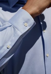 Helles blau karierter Hemdärmel mit strukturierter Oberfläche, zwei weiße Knöpfe zeigend, naher Blick auf den Stoff und die Details.