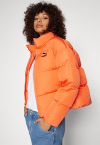 Giacca imbottita arancione brillante con colletto alto, chiusura con zip e un logo nero. Presenta un design cropped e una texture morbida e liscia.