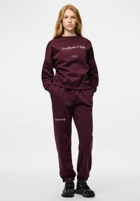 Burgundski pulover in športne hlače iz mehke tkanine z belim napisom "Aesthetic Club". Pulover ima okrogel ovratnik; hlače pa elastične manšete.