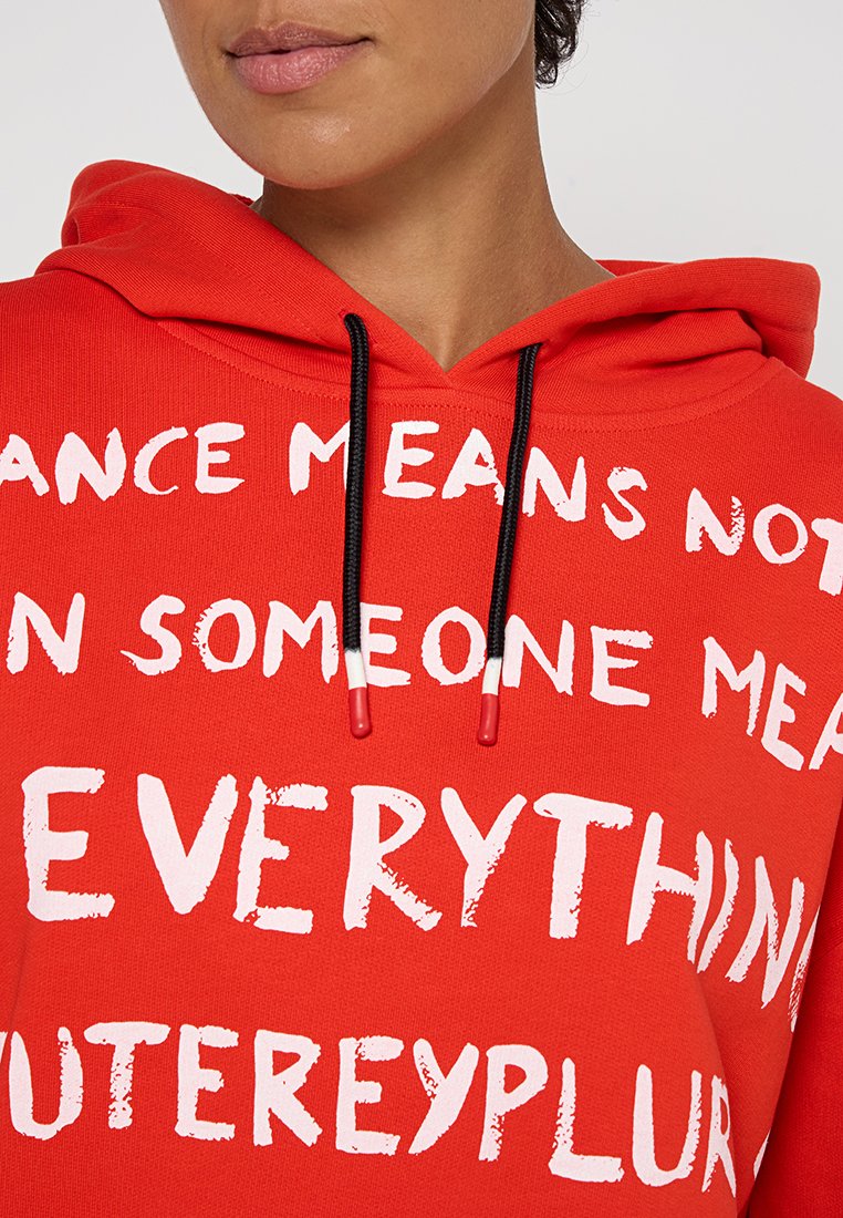 Czerwona bluza z kapturem z białym napisem w różnych czcionkach, czarnymi sznurkami ściągającymi z czerwonymi końcówkami oraz miękką bawełnianą fakturą. Napis zawiera motywacyjne hasła.