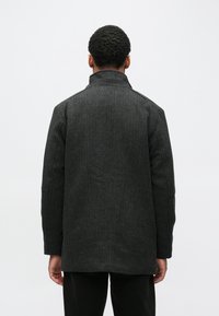 Temno sivi volneni plašč s strukturo, visokim ovratnikom in teksturiranim materialom. Ima raven kroj in minimalistične detajle.