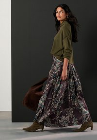 Olivově zelený svetr, dlouhá květinová plisovaná sukně v tmavě fialové s modrými akcenty, hnědá taška a boty na podpatku v barvě taupe. Hladké textury.