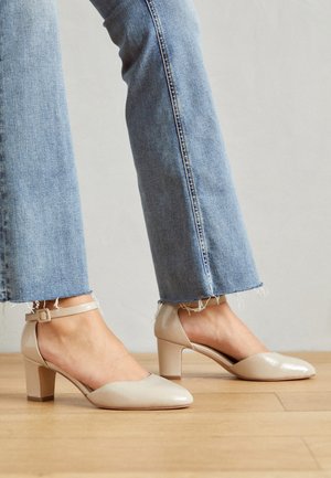 Beige Lackleder-Pumps mit quadratischer Spitze, Knöchelriemen und mittelhohem Blockabsatz. Kombiniert mit blauem Jeansstoff mit ausgefranstem Saum.