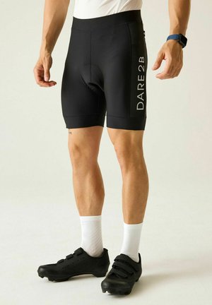 Uomo che indossa pantaloncini da ciclismo neri con logo "DARE 2 B", calze bianche, scarpe da ciclismo nere e un orologio da polso blu su sfondo uniforme.