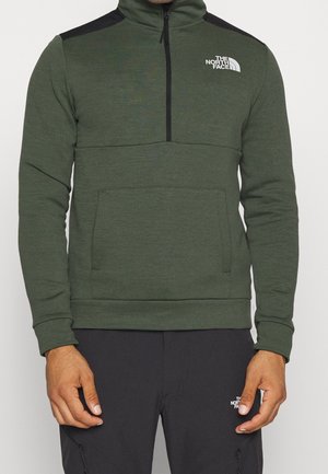 Mann trägt einen grünen Fleece-Halbzip-Pullover mit schwarzen Schulterpatches und schwarze Hose, beide mit The North Face-Logos.