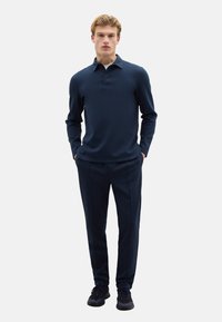 Dunkelblaues Polohemd mit langen Ärmeln, Kragen und drei Knöpfen, kombiniert mit maßgeschneiderten dunkelblauen Hosen und dunklen Turnschuhen, die eine taillierte Silhouette zeigen.