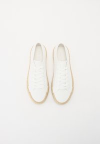 White canvas sneakers with beige woven jute soles, six eyelets for laces, and a smooth finish. Minimalist design, flat shape, and no visible logos.