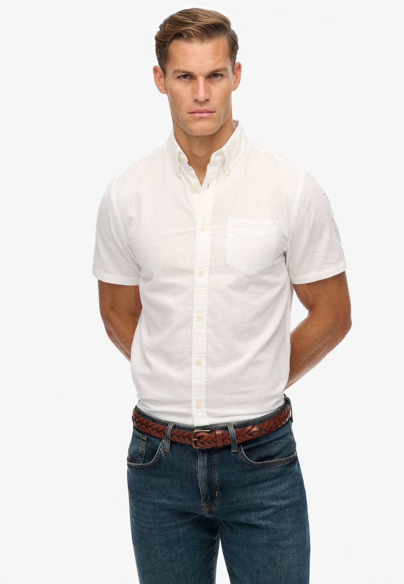 Camicia bianca a maniche corte con bottone e taschino, abbinata a jeans in denim blu e una cintura intrecciata marrone.