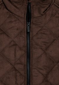 Bruine quilted stof met een centrale zwarte ritssluiting en metalen ritsluiting aan de kraag van een gewatteerde jas of vest.