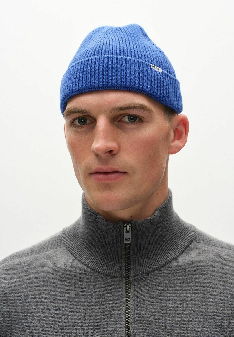 Blå stickad mössa med ribbad textur, prydd med ett litet varumärkesmärke. Tillsammans med en grå zip-up-tröja med hög krage.