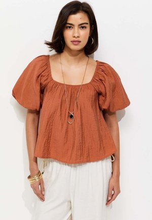 Vrouw draagt een roestoranje top met pofmouwen, witte broek, gouden armbanden, grote oorringen en een lange hangerketting, staand tegen een witte achtergrond.