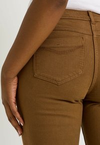 Jean en denim marron avec une coupe ajustée, doté d'une poche arrière classique et de coutures discrètes. Texture lisse et forme cintrée.