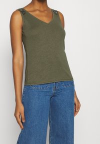 Top sin mangas verde oliva con escote en V, que presenta un embellishment de cuentas en los hombros. Tejido suave y diseño ajustado, combinado con jeans azules.