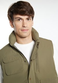 Khaki-Weste mit hohem Kragen, Taschen vorne und Reißverschluss, kombiniert mit einem weißen Rundhals-T-Shirt darunter. Glattes, leichtes Material.