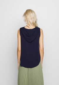 Haut sans manches bleu marine avec une capuche, texture lisse et ourlet arrondi, associé à une jupe verte plissée. Vue arrière.