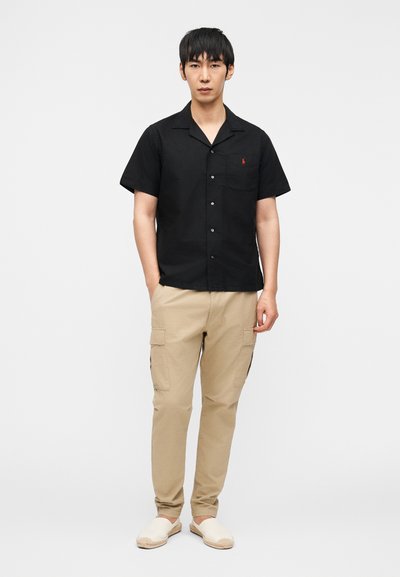 Άνδρας με μαύρο κοντομάνικο πουκάμισο με κουμπιά, μπεζ παντελόνι cargo και κρεμ παπούτσια χωρίς κορδόνια, στέκεται μπροστά σε λευκό φόντο.