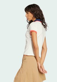 Biela polokošeľa s ružovými a námorníckymi akcentmi, krátkymi rukávmi a priliehajúcim strihom. Model ju kombinuje s béžovou plisovanou sukňou.