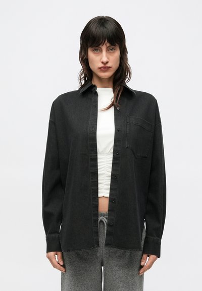 Schwarzes Denimhemd mit durchgeknöpftem Vorderteil, einer Brusttasche und lockerer Passform. Das Modell trägt ein weißes Oberteil und graue Hosen. Glatte Textur.