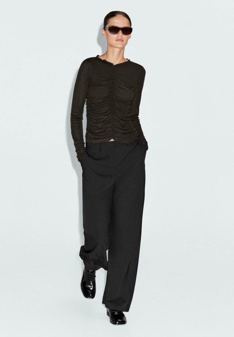 Camicia scura marrone a maniche lunghe con dettagli arricciati, abbinata a pantaloni neri a vita alta e scarpe stringate nere. Gli occhiali da sole completano il look.