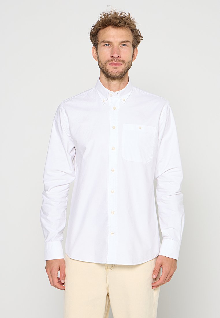 Man met krullend haar in een knoopshirt met lange mouwen in het wit, met een borstzak en beige broek, staande voor een effen achtergrond.