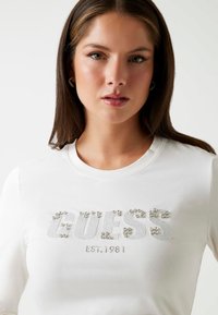 Witte langekorte mouw t-shirt met een met rhinestones versierde "GUESS" logo en de tekst "EST. 1981", met een ronde halslijn en een zachte stoftextuur.