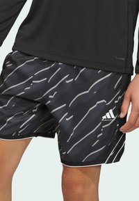 Mustat urheilushortsit, joissa on harmaa abstrakti kuvio, valkoisia yksityiskohtia ja pieni logo, valmistettu kevyestä ja hengittävästä materiaalista.