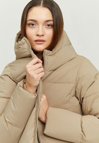 Beige puffer jacket with a high collar and zip closure. Features quilted texture and tonal branding on the chest. Soft, insulated material.
