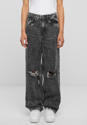 Person wearing black acid-wash wide-leg jeans with large ripped holes at the knees and white sneakers, standing against a plain background.