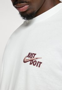 Hvid bomulds t-shirt med et maroon "Just Do It" logo og en swoosh grafik, rund halsudskæring og korte ærmer.