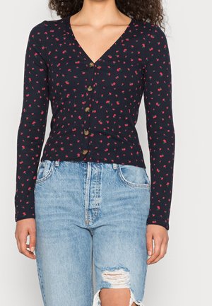Navy ribbed long-sleeve cardigan with a V-neck, featuring small red floral patterns and brown buttons down the front. Worn with distressed denim.