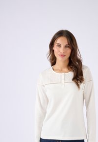 Langarmige crème top met een ronde halslijn, een decoratieve kantpaneel over de borst en drie knopen aan de voorkant. Zacht, ademend materiaal.