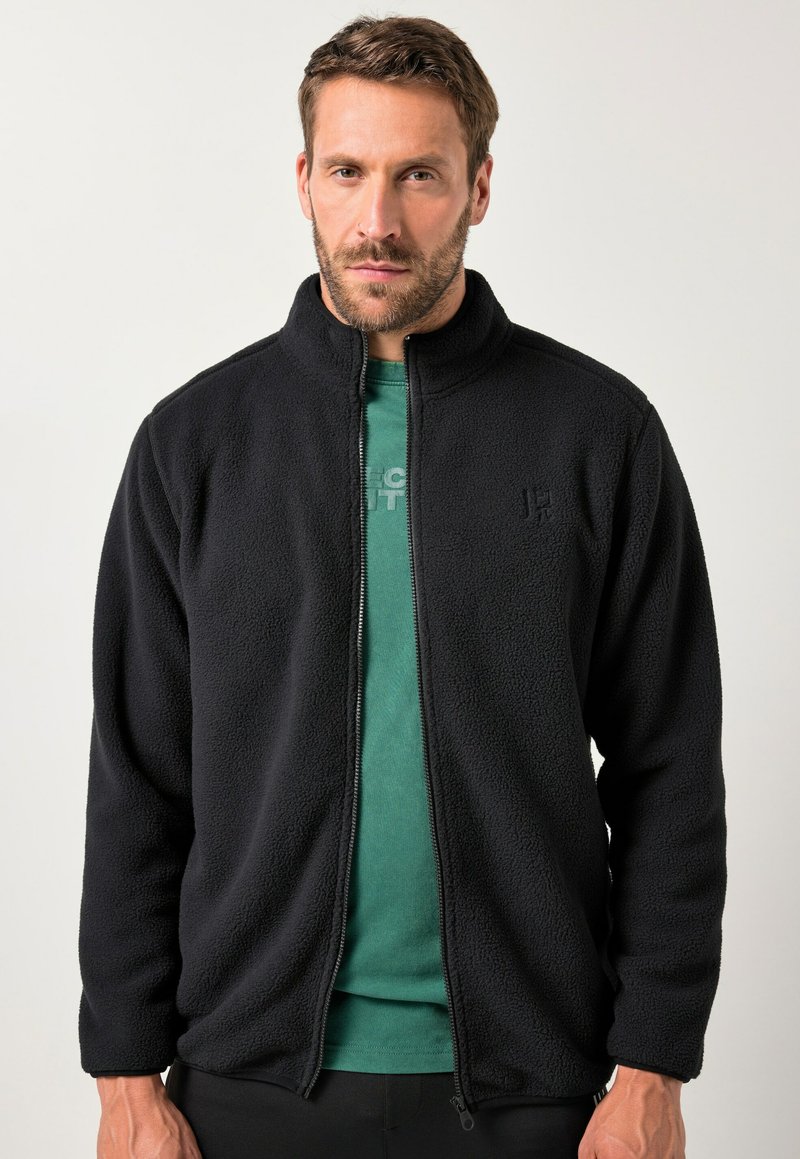 Chaqueta negra de forro polar con cuello alto, cremallera frontal y tejido texturizado. Se lleva sobre una camisa verde, con un logo sutil en el pecho.