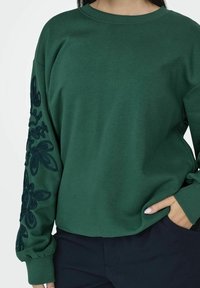 Sudadera verde de corte relajado, con un patrón de bordado floral texturizado en la manga izquierda y puños acanalados.