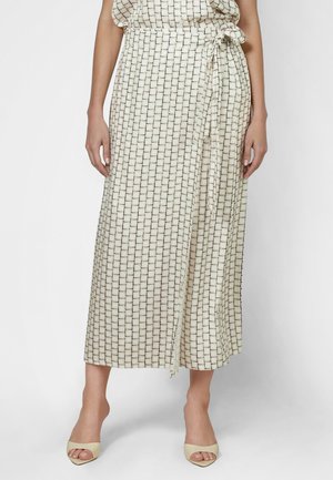 Frau trägt einen beigen Wickel-Midirock mit schwarzem Ziegelmuster und beigefarbene offene High Heels, steht vor einem einfarbigen Hintergrund.