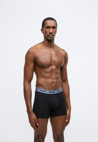 Cuecas boxer pretas para homem com uma cintura elástica azul clara que apresenta o nome da marca "HUGO". O tecido parece liso e ajustado.