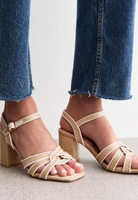Beige strappy heeled sandals with a square toe, woven texture, and sturdy block heel, paired with frayed denim jeans.