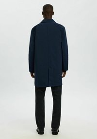 Marineblauer, taillierter Mantel mit geradem Schnitt, langen Ärmeln und einem Rückenschlitz. Getragen über dunklen Jeans und schwarzen Schuhen. Glatte Textur.