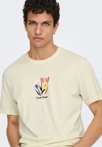 Ανοιχτό κίτρινο βαμβακερό t-shirt με λουλουδάτο σχέδιο που απεικονίζει τουλίπες σε ροζ, κόκκινο, μπλε και κίτρινο, συνοδευόμενο από το κείμενο "Jardin Parisien".