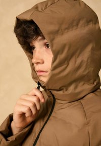 Veste à capuche matelassée marron avec un col haut, dotée d'une fermeture éclair noire et d'une texture lisse, légèrement brillante. Gros plan d'une personne la portant.